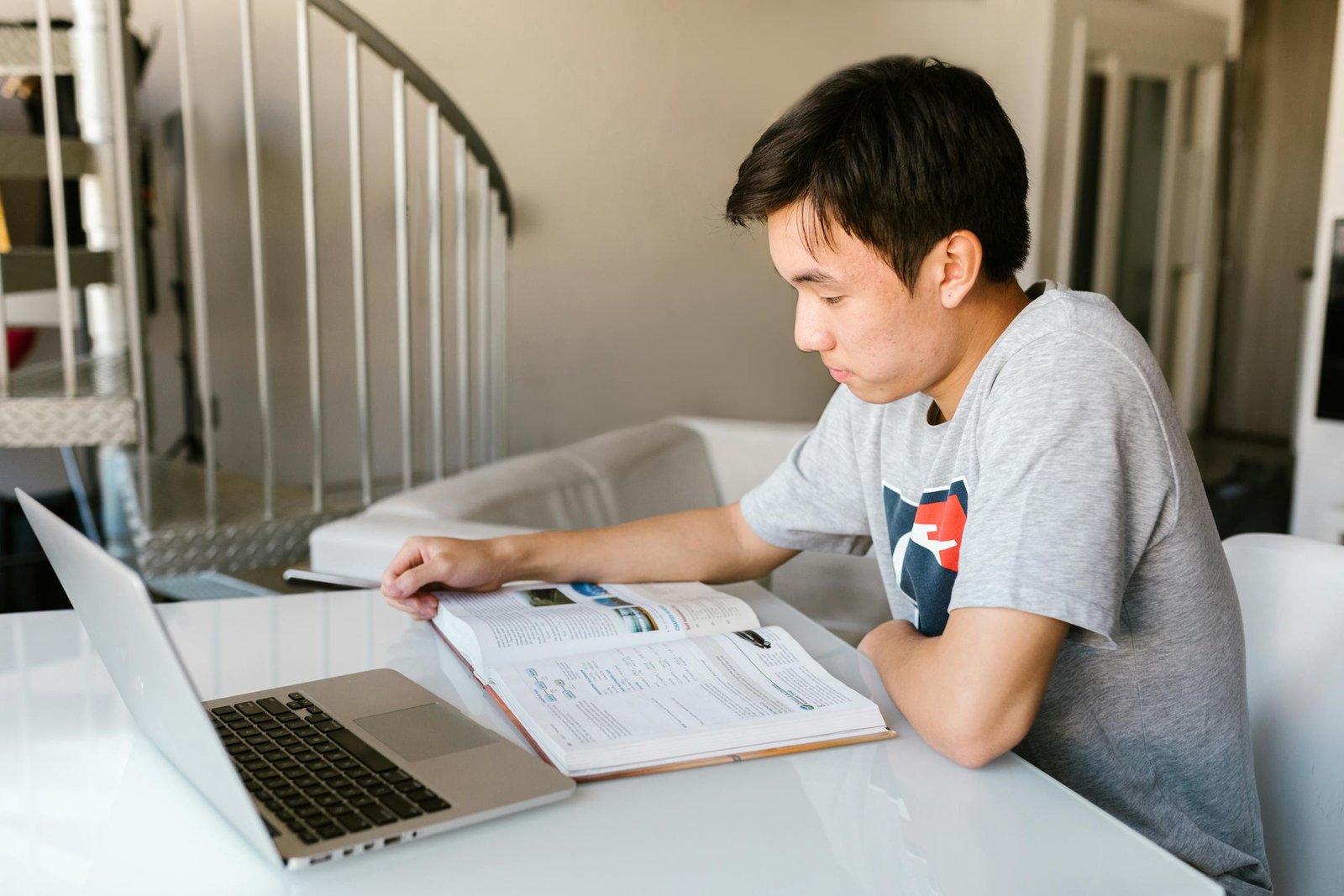 Teenage boy focuses on homework using a laptop and textbook indoors.
